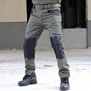 Person wearing gray and navy tactical pants with black boots on a concrete floor.